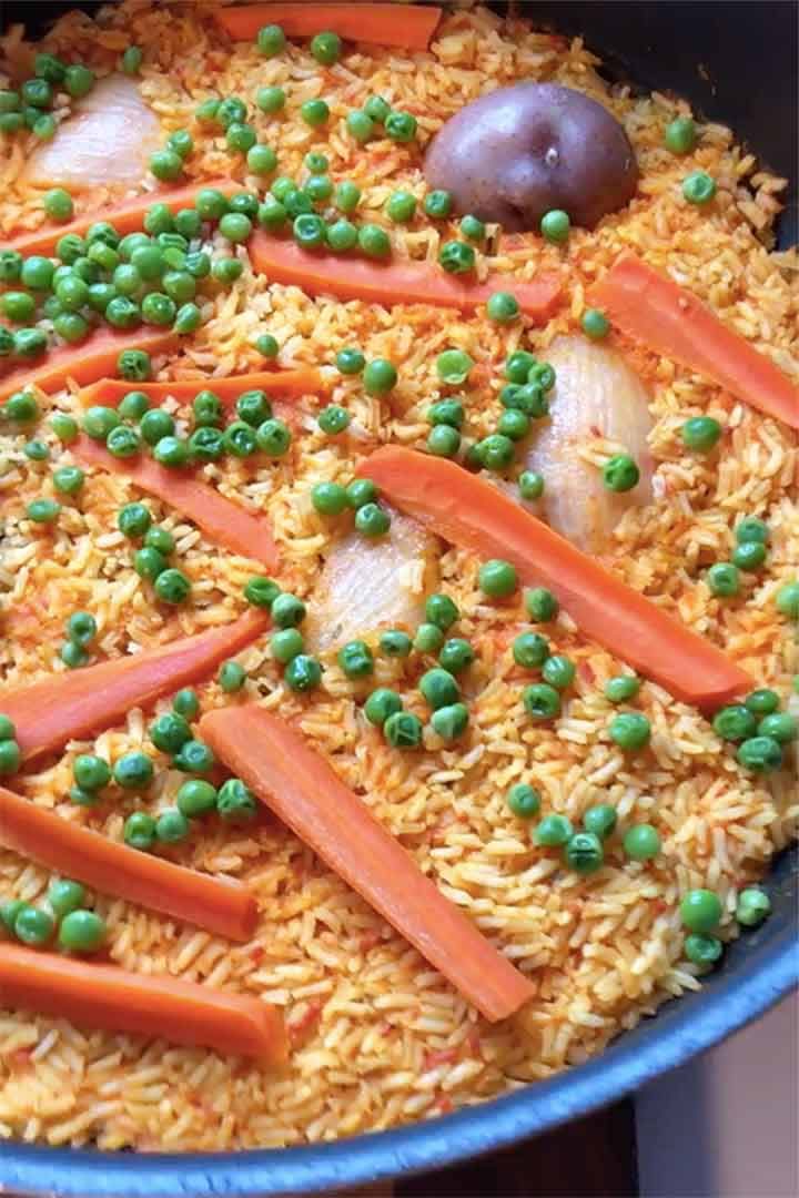 Close-up image of cooked rice with peas sliced carrots and red potato in large pot.