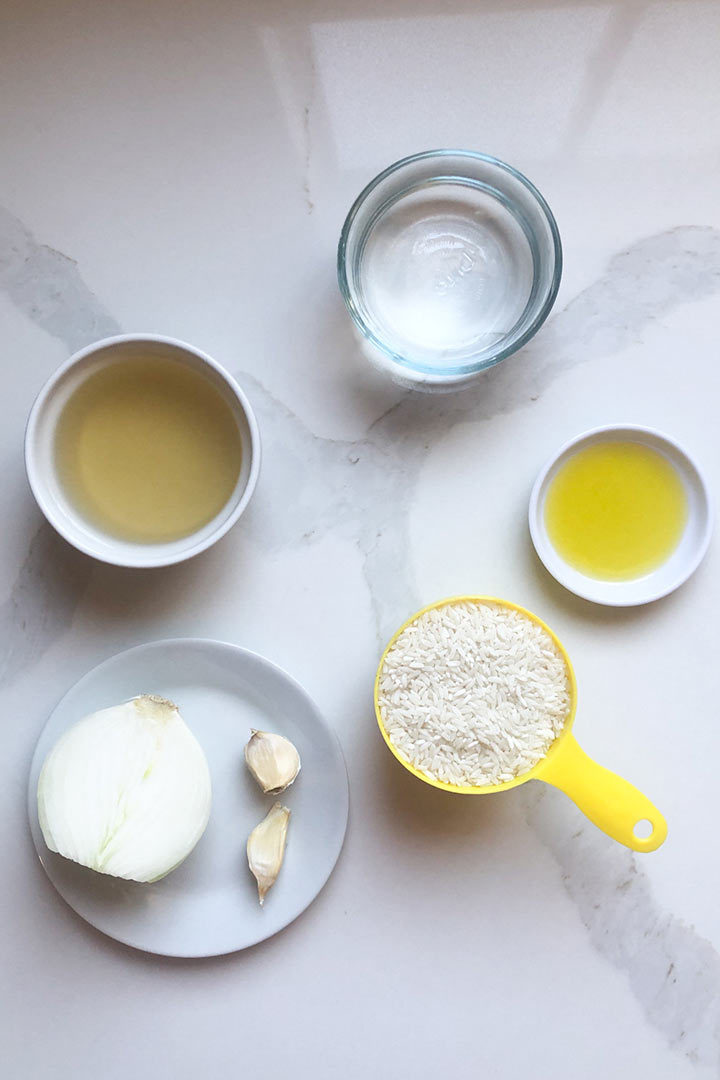 ingredients for recipe on a counter: water, chicken broth, oil, onion halve, garlic cloves, and long grain rice.
