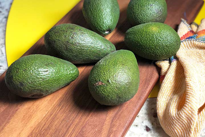 six fresh and ripe avocados on a dark wood surface.