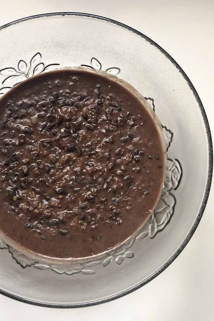 Off centered clear glass bowl with etched flowers filled with Refried Black Beans on white surface.