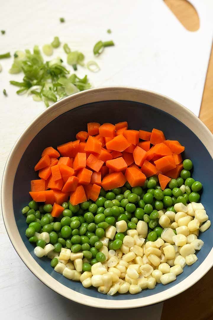 Vegetable ingredients in a round bowl and slivered green onions in background on white surface.