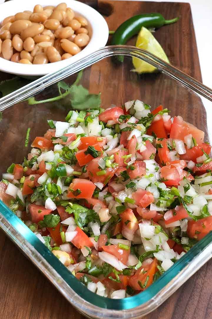 Essential Pico de Gallo Recipe in a glass dish with a small bowl of pinto beans and serrano pepper in background.