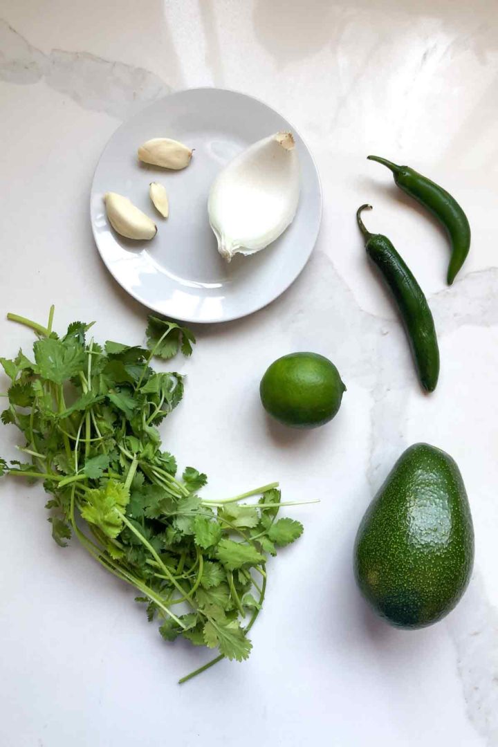 ingredients for Spicy Avocado salsa for tacos, fresh cilantro, serrano, lime, onion, and garlic on white surface.