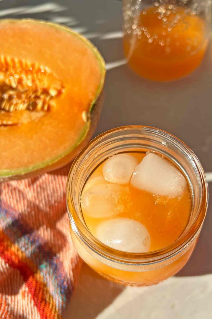 Agua de Melon Drink Recipe on ice in glass with halved fresh melon and red knit cloth on a patio.