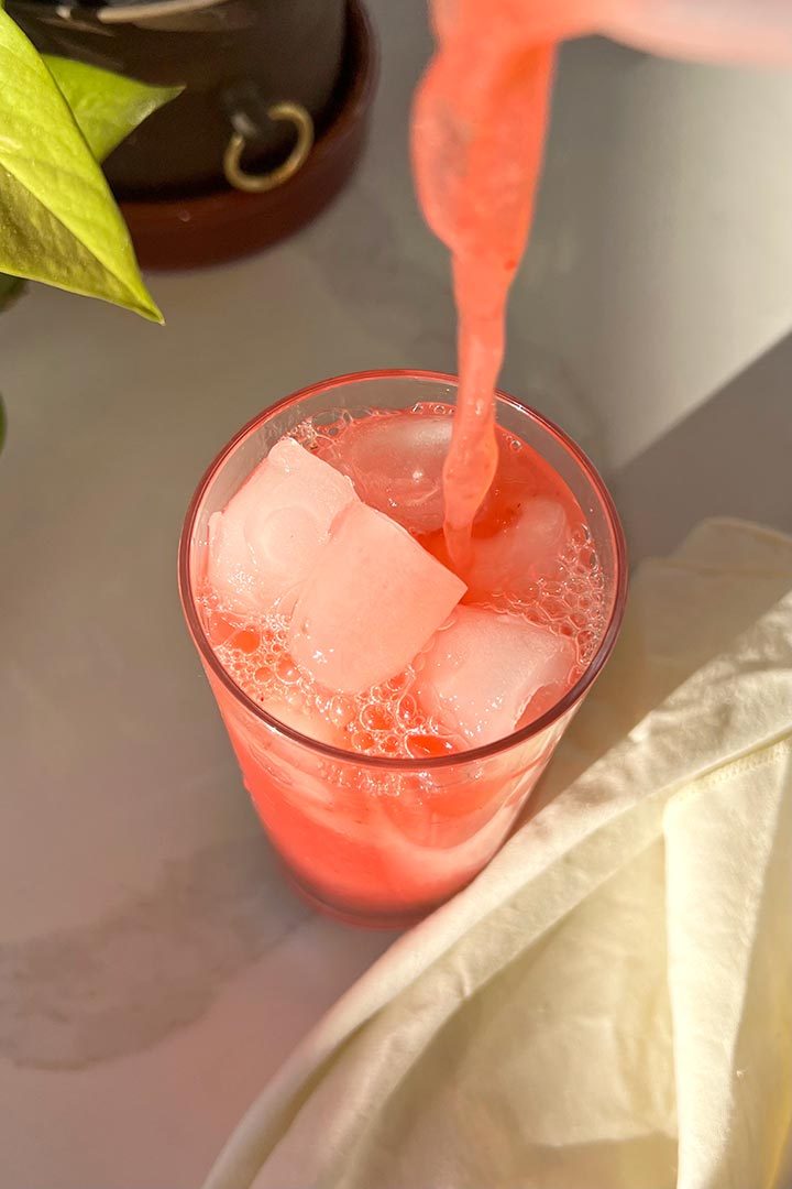 Pouring Strawberry water over ice into cup on white surface.