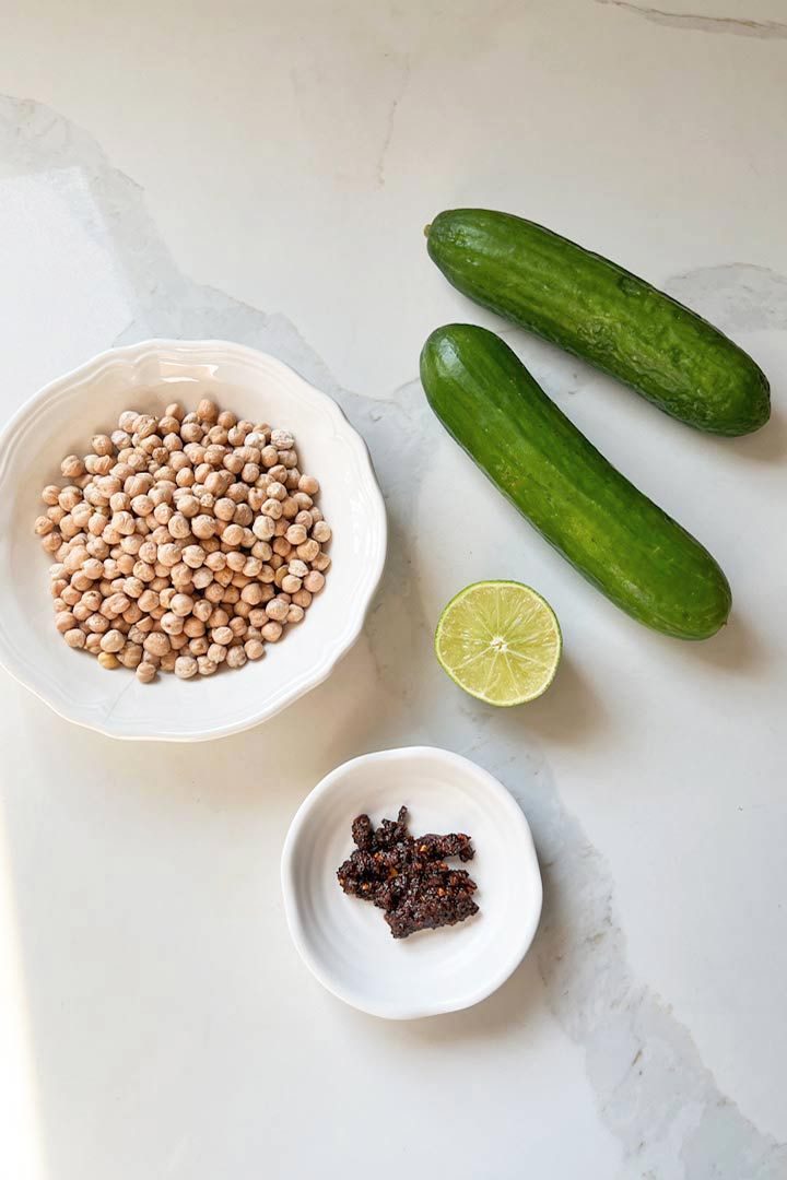 Ingredients to make Garbanzo Snack; chickpeas, cucumbers, lime, salsa macha, on white surface.