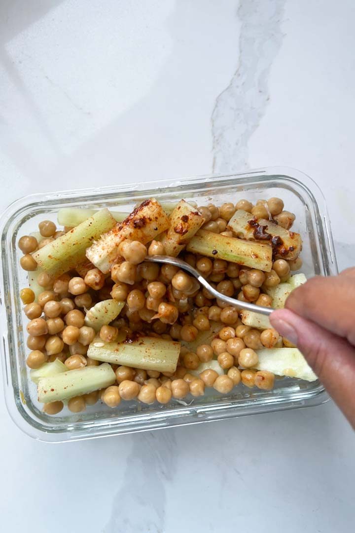 Brown hand holding spoon and mixing ingredients for cooked healthy Chickpea Snack on white surface.