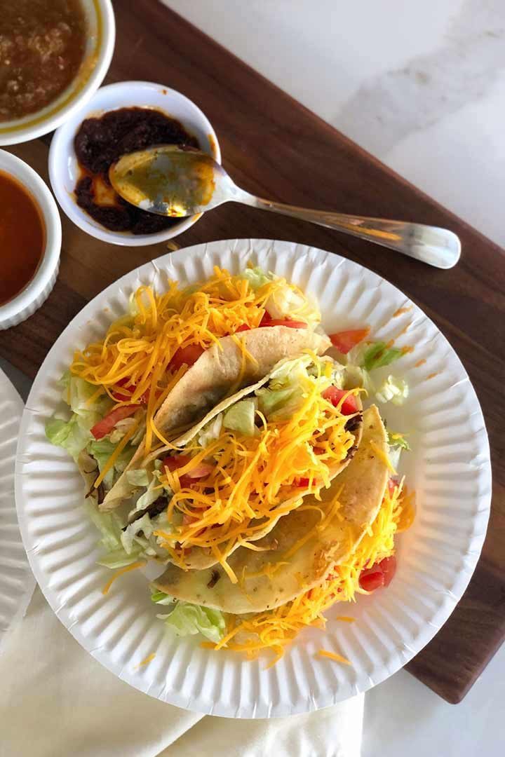 Three hard shell tacos recipe Final on a white paper plate with salsa beside it on wood board.