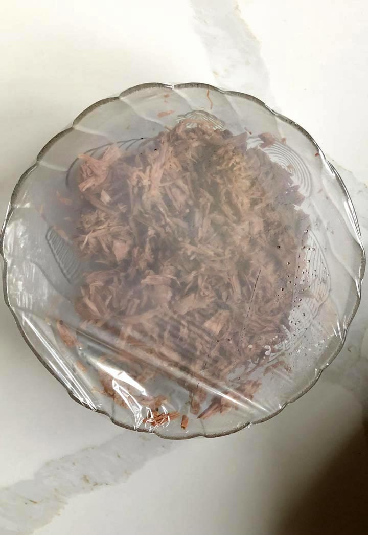 Covered shredded beef in round glass bowl to seal moisture on a white surface.