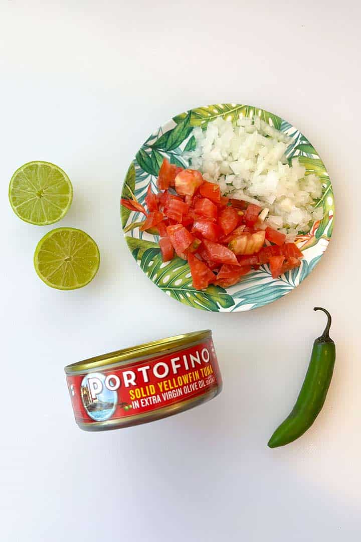 Ingredients for tuna salad mexican style on a white surface; lime, onion, tomato, tuna, and peppers.