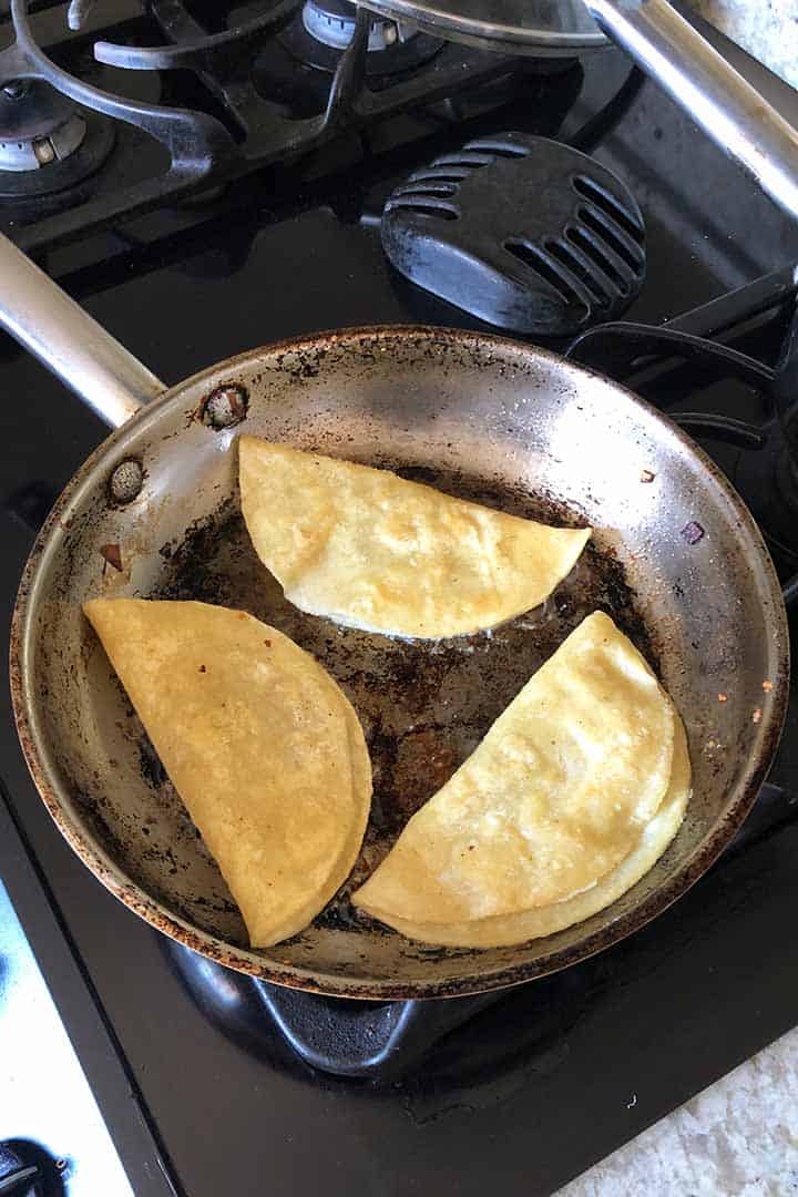 frying three potato tacos until crispy in a round pan on a black stove top.