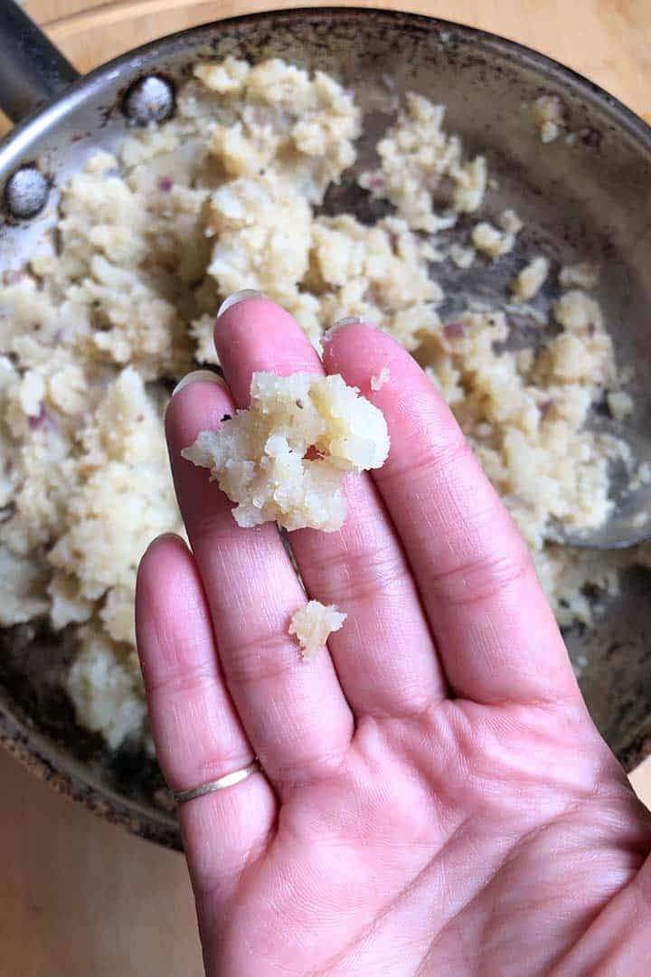 The potato for tacos on fingertips to share the cooked texture with pan of potato in background.