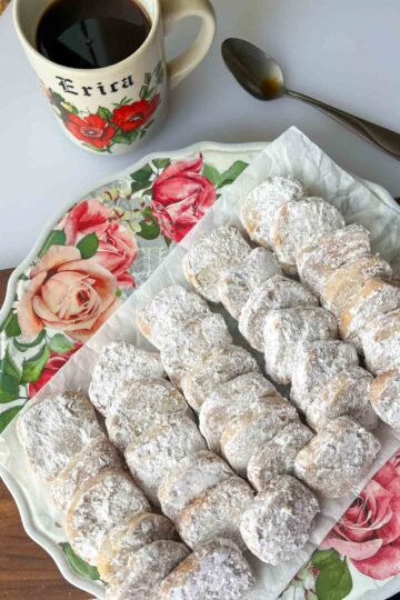 A platter of Mexican Wedding Cookies on floral plate with black coffee in a decorative mug above it.