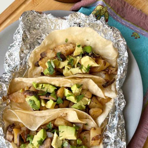 Cozy Mushroom and Potato tacos with diced avocado and cilantro garnish nestled in a piece of aluminum foil to keep them warm.