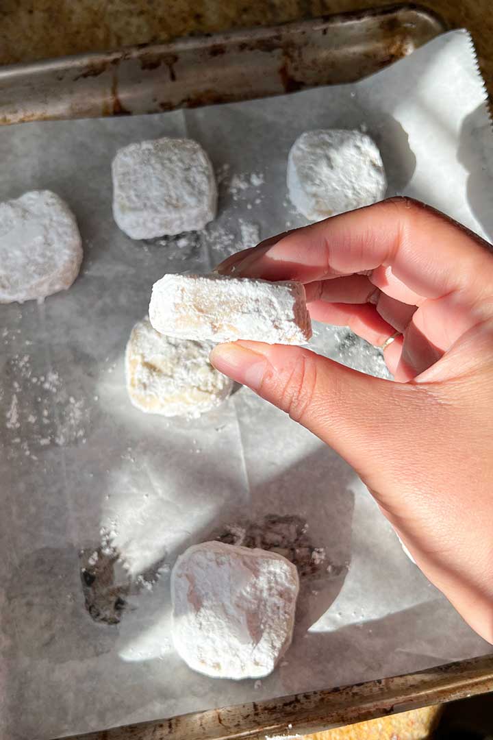Hand holding up Mexican Wedding Cookie once cooled with baking sheet in background.
